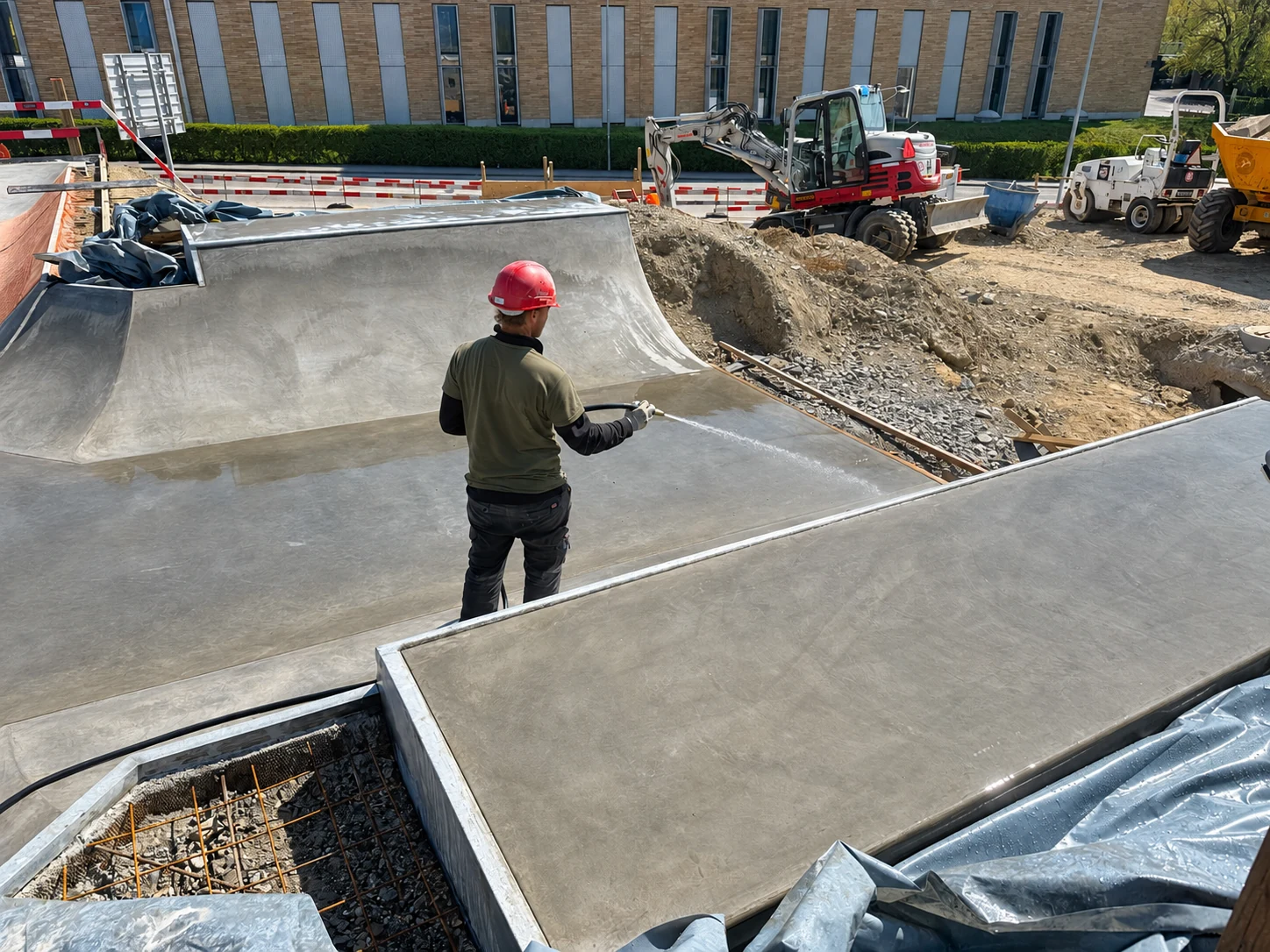 Baustellenfoto eines Beton-Skateparks mit Arbeiter, Minibagger und frischen Betonflächen.