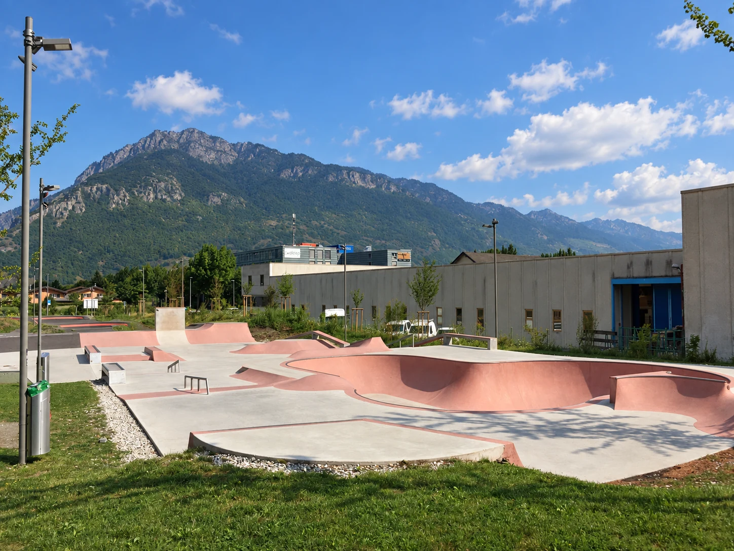 Beton-Skatepark mit rosafarbenen Bowls, Street-Elementen und Bergpanorama in einer sonnigen Gesamtansicht ohne Personen.