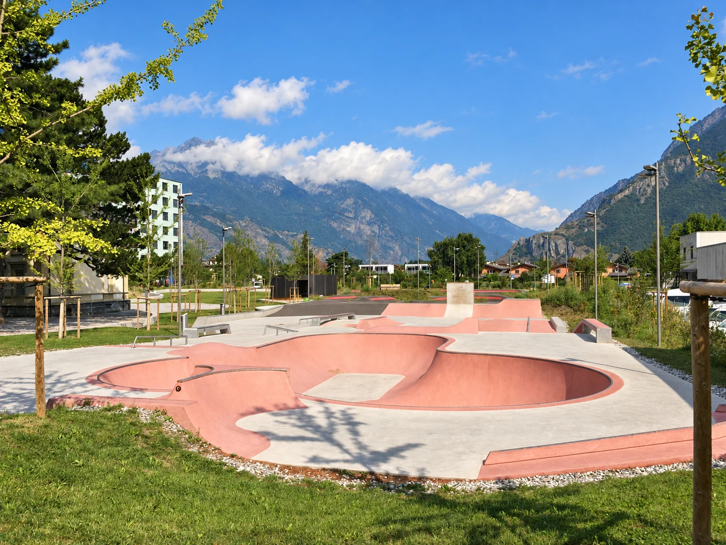 Weitläufiger Beton-Skatepark mit rosafarbener Bowl, Rampen und Rails in grüner Parklandschaft vor Bergen.