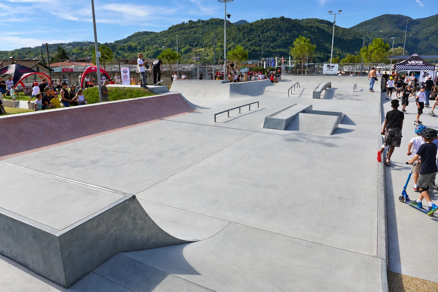 Weitansicht eines Beton-Skateparks mit Street-Area, Zuschauern und grüner Bergkulisse.