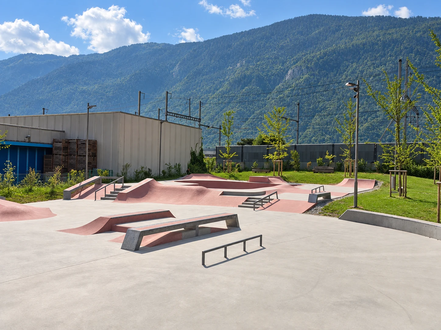 Leerer Beton-Skatepark mit Rails, Ledges, Treppen und rosafarbenen Rampen in sonniger Weitwinkelansicht.