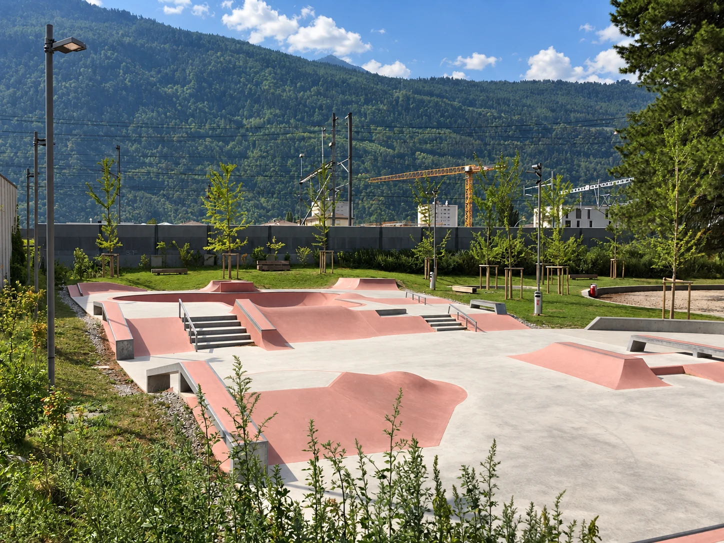 Sonniger, menschenleerer Beton-Skatepark mit rosafarbenen Street-Rampen, Treppen und Rails vor bewaldetem Berg.
