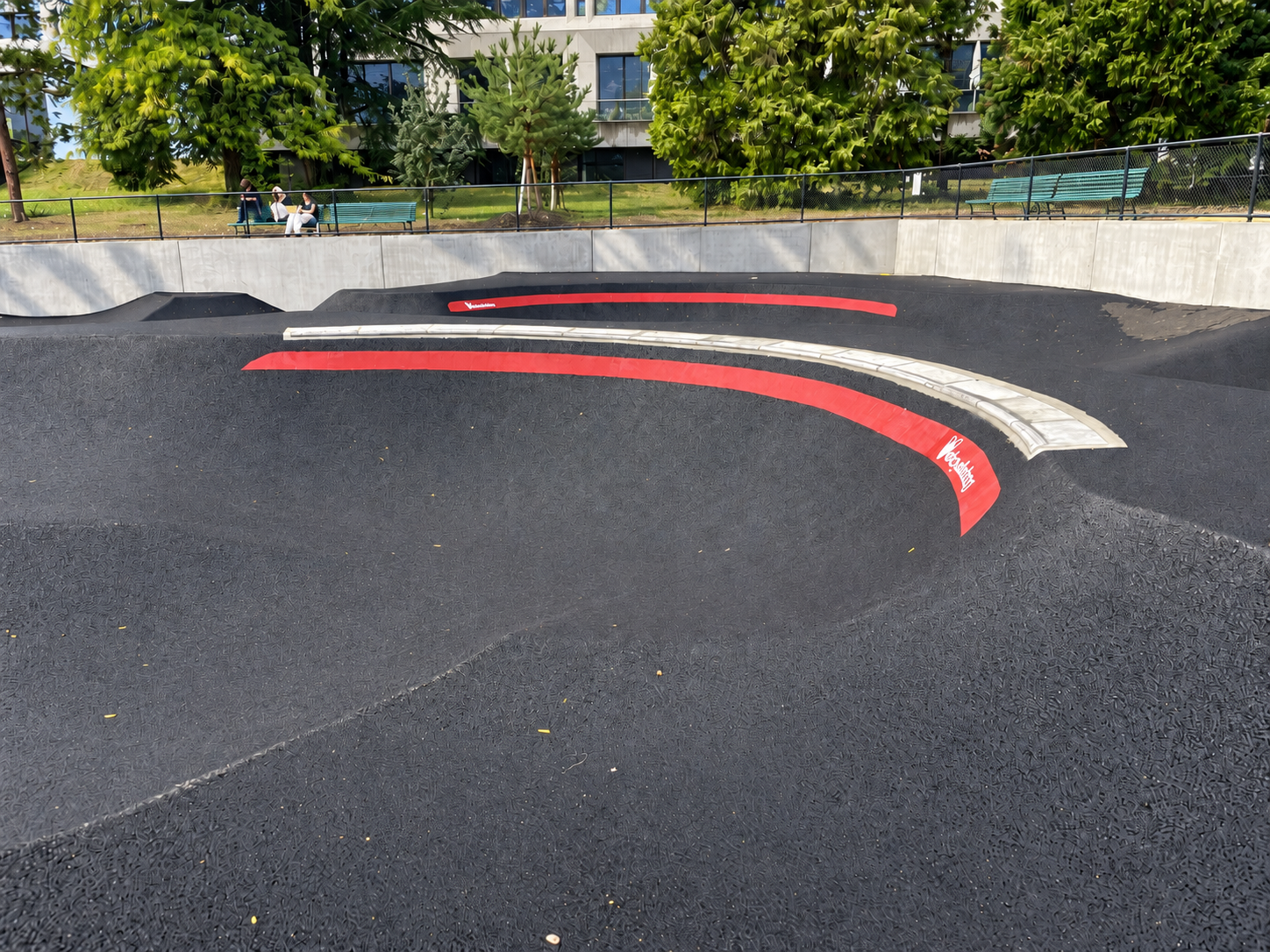 Kurvendetail des Asphalt-Pumptracks im Parc des Franchises in Genf mit roter Markierung und heller Kante, dahinter grüne Sitzbänke.