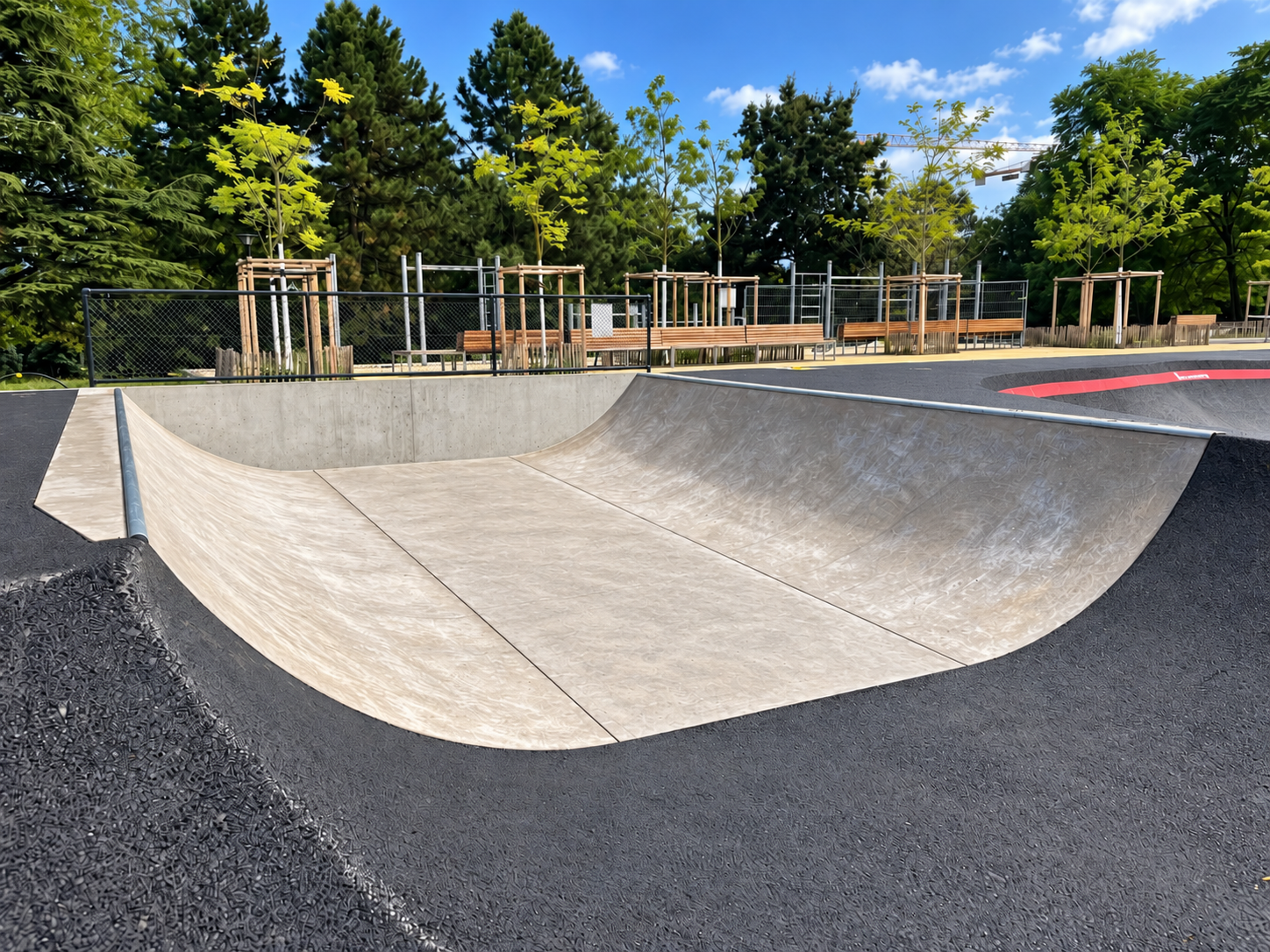 Beton-Miniramp im Parc des Franchises in Genf mit geschwungenen Übergängen und Metallcopings, dahinter Holzbänke und junge Bäume.